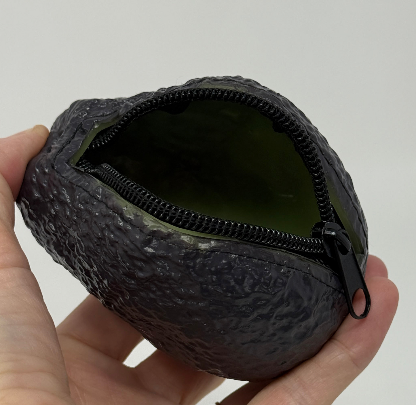 Close up image of a hand displaying a coin purse that is a realistic replica of an avocado with a bumpy green exterior. The coin purse has a short zipper to keep the contents safe.